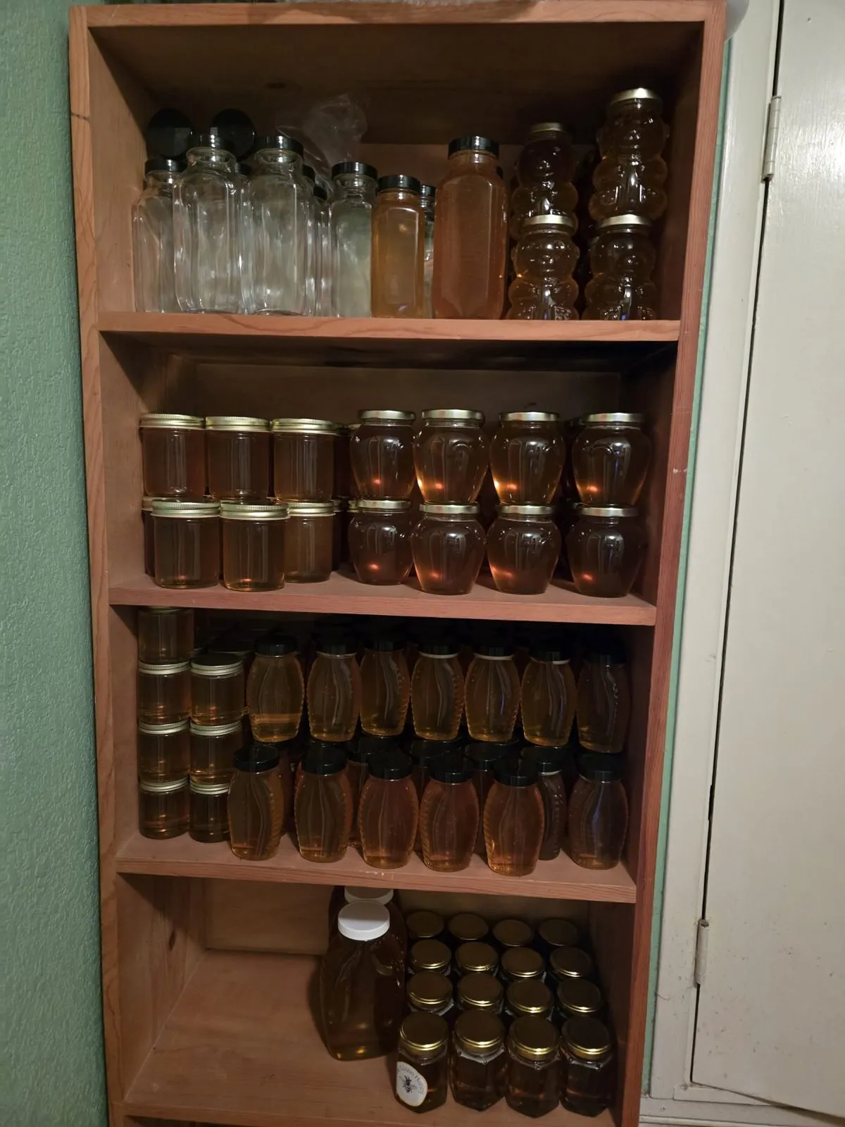 Wooden bookcase of the 2026 pack: queenline jars with gold lids, ribbed honey bottles, and empty glass waiting to be filled