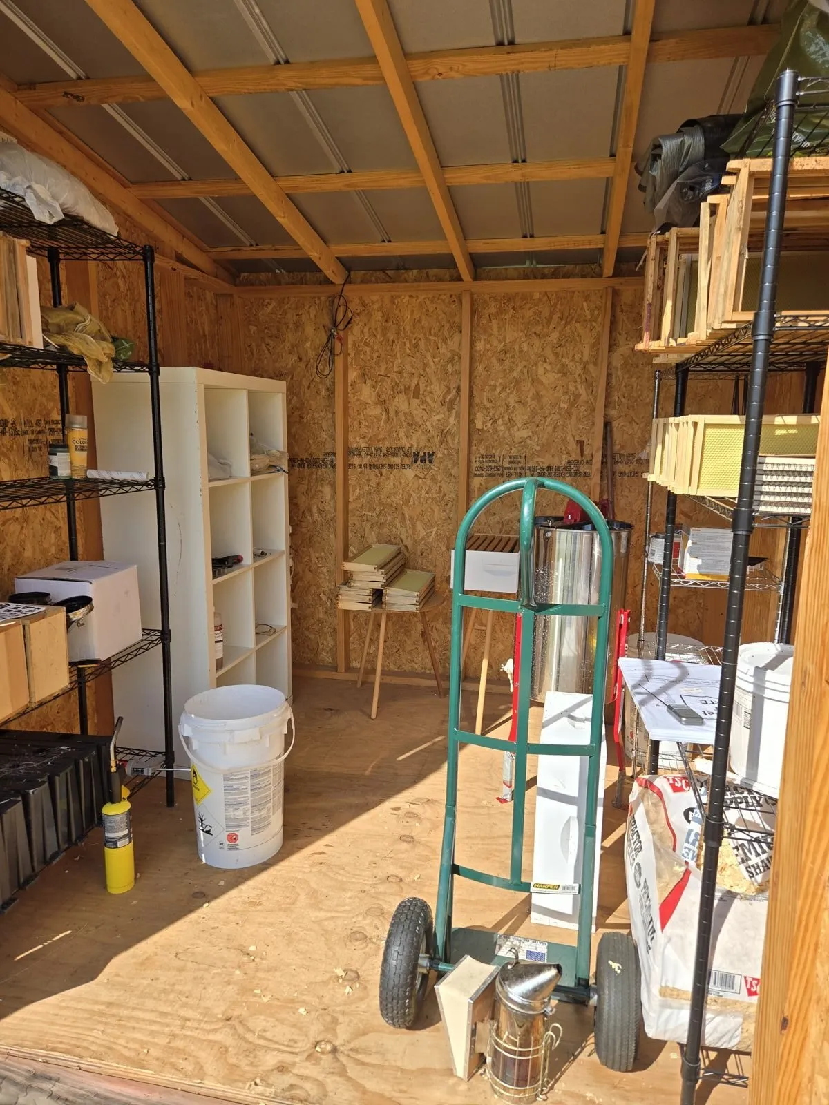 Interior of the honey house: plywood walls, metal extractor, green hand truck, smoker, and stacked wooden frames